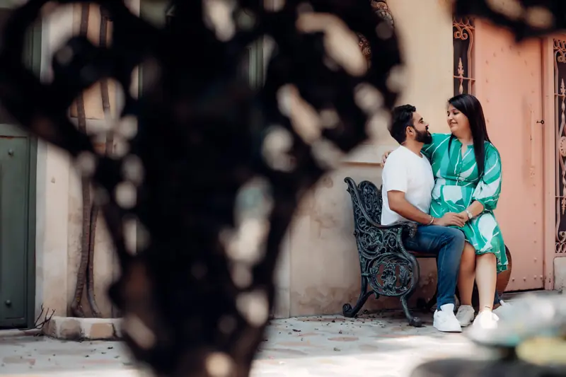 Couple seated on a garden bench, framed through foreground foliage during a pre-wedding shoot